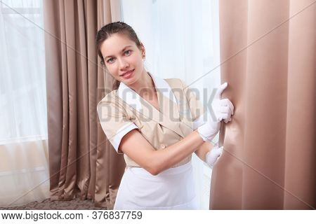 A Maid In White Gloves And Uniform Smiles And Adjusts The Curtains In A Hotel Room Or House. The Con