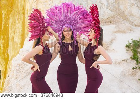 Three Girls In Carnival Costumes Pose And Smile Against The Background Of A Rock