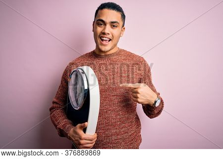 Young brazilian man doing diet to lose weigth holding scale over isolated pink background very happy pointing with hand and finger