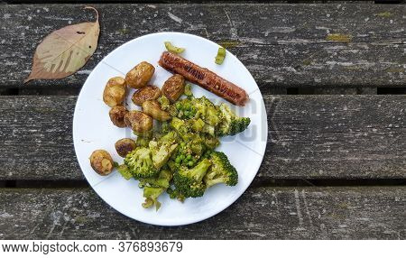 Vegan Meal With Oven Baked Potatos, Plant Based Sausace, Broccoli, Peas And Pesto On A White Plate. 