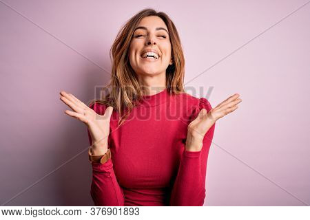 Young beautiful brunette woman wearing casual sweater stnding over pink background celebrating mad and crazy for success with arms raised and closed eyes screaming excited. Winner concept