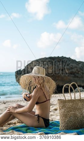Beautiful Young Woman On The Beach Applying Sunblack While Sitting On Her Towel And Wearing Big Stra