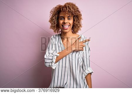 Beautiful african american woman with curly hair wearing striped t-shirt over pink background cheerful with a smile of face pointing with hand and finger up to the side with happy and natural expressi