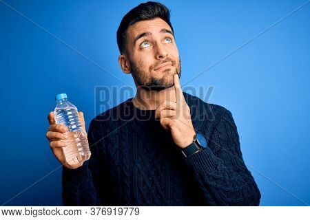 Young handsome man drinking bottle of water to refeshment over blue background serious face thinking about question, very confused idea
