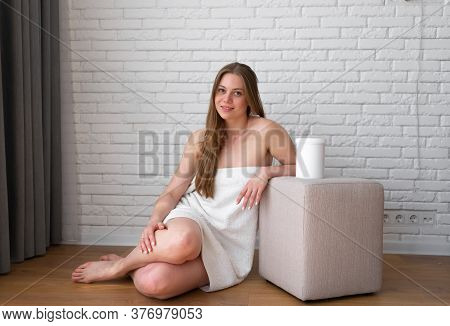 Positive Smiling Woman Wrapped In Bathtowel Sits By Poof Chair With Sugaring Paste Jar On It.