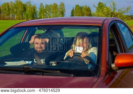 A Woman Holds A Drivers License In Her Hands. A Driving Instructor Sits Next To Him And Holds His Ha