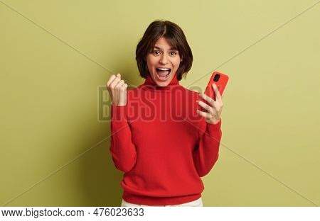 Cheerful Young Female With Short Hair In Red Turtleneck Standing Near Green Background With Smartpho