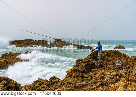 Oualidia, Morocco - April 08, 2023: View Of A Fisherman, And Waves Crashing On The Rocky Beach, In T