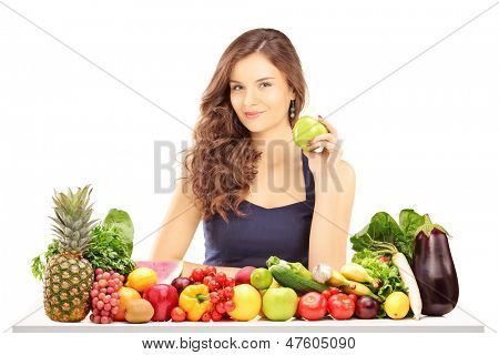 Giovane sorridente femmina tenendo una mela verde e in posa dietro un tavolo pieno di frutta e vegatebles i
