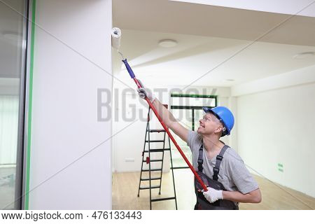 Portrait Of Smiling Decorator In Helmet Painting Wall With Roller Brush. Painting, Renovation And Ho