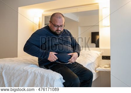 Man Suffering From Extra Weight Touching Abdomen Sitting On Bed In Bedroom. Overweight Bearded Balde