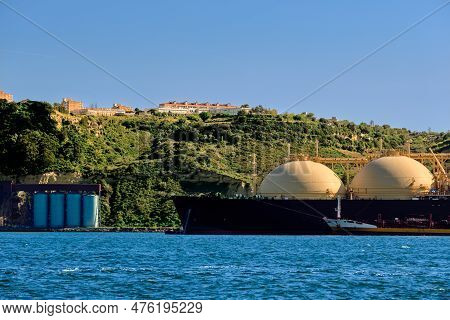 LNG or liquified natural gas tanker moored by industrial pier and green hill on sunny day. Alternative gas supply, commercial freight, energy crisis, regasification, diversification, fuel and energy generation.
