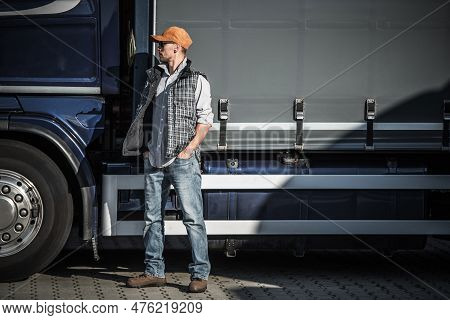 Caucasian Semi Truck Driver In His 40s In Front Of His Tractor Trailer. Truck Stop Break.