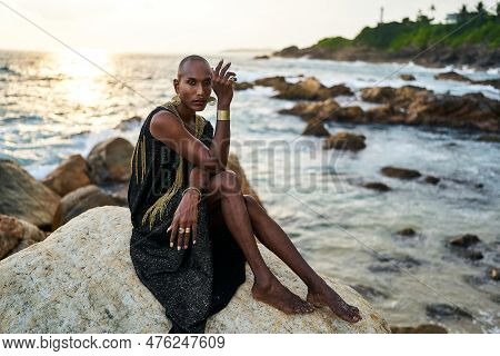 Non-binary Black Person In Luxury Dress Sits On Rocks In Ocean. Trans Ethnic Fashion Model Wearing J