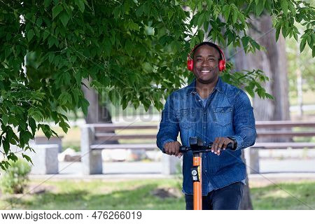 Cute Black Student Enjoys Nature Riding An Electric Scooter In A Large Green Park On A Sunny Day. Mo