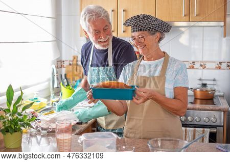Happy Senior Couple In The Home Kitchen While Wife Holds A Freshly Baked Homemade Plumcake And Husba