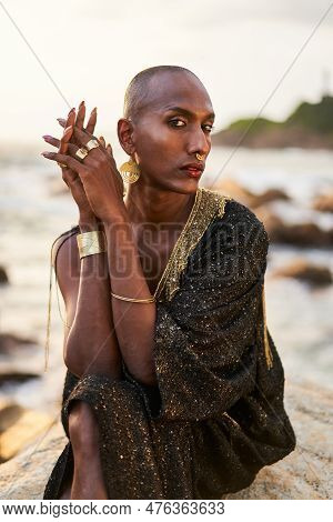 Non-binary Black Person In Luxury Dress Sits On Rocks In Ocean. Trans Ethnic Fashion Model Wearing J
