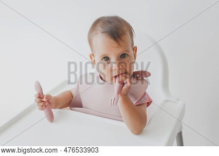 Close-up Portrait Of A Cute Baby Girl Infanta 8 Months Old, Sitting On A Highchair In A Baby Apron W