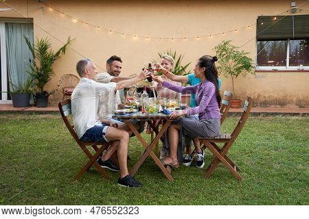 Group Of Middle-aged Friends Toast With Wine Gathered Around A Garden Table On A Summer Evening To S
