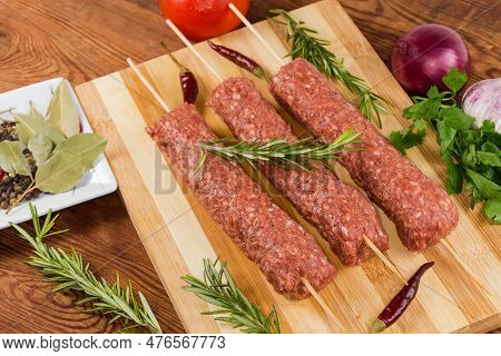 Several Raw Lula Kebab Skewered On Wooden Skewers On The Wooden Cutting Board Among The Some Vegetab