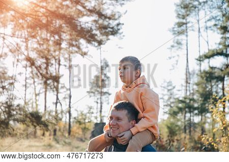 Dad And Son On A Walk In The Forest In Sunny Weather. Family Leisure. The Father Carries His Son On 