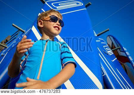 Blond Boy In A Blue Suit And Sunglasses Cleans His Hands Of Sand Against The Background Of Sports Eq