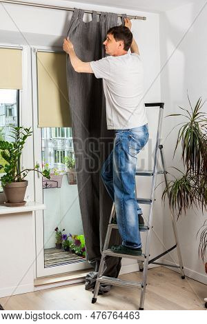A Man Stands On A Stepladder And Hangs Gray Curtains On A Curtain Rod In An Apartment, Spring Cleani