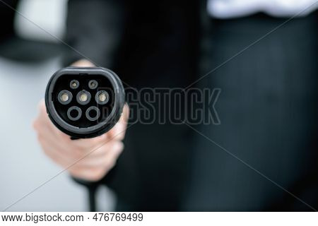 Focus Closeup Hand Holding Ev Plug For Electric Vehicle Pointing At Camera With Charging Station At 