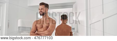 Positive Man With Towel And Crossed Arms Leans Onto Cabinet Near Mirrror In Bathroom