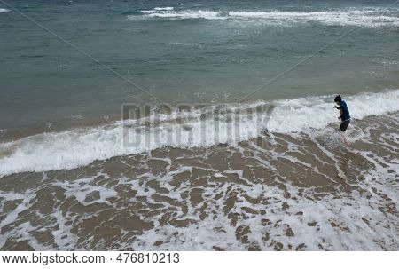 Drone Aerial Of Young Boy In Shorts Standing On The Summer Beach And Looking At The Stormy Spindrift