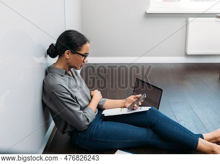 Young Female Entrepreneur In Thought Sitting On The Floor In The Kitchen With A Laptop On Her Legs