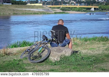 Man With Bicycle Sitting On O Big Stone Looking At A River Blue Water And Bike Lying On Green Grass 