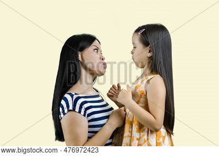 Side View Of Mother And Her Little Girl Are Making Faces To Each Other. Isolated On Pastel Yellow.