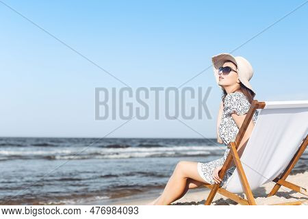 Happy Brunette Woman Wearing Sunglasses And Hat Relaxing On A Wooden Deck Chair At The Ocean Beach
