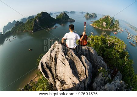 A young traveler man sit on the top of mountain with his guitar in Halong bay and enjoy the beauty of seascape.