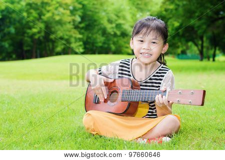 Asian Little Girl Sitting On Grass And Play Ukulele In Park
