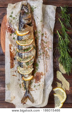 Whole baked mackerel or scomber fish with lemon and greens on paper dark wooden background top view