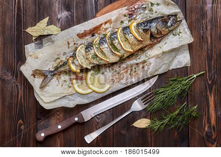 Whole baked mackerel or scomber fish with lemon and greens on paper dark wooden background top view