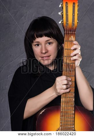 beautiful fashionable lady wearing a gothic black dress with high collar, poses with an acoustic guitar on a grey background