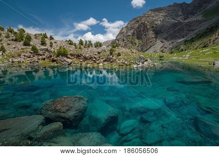 Blue and transparent lake in Fann Mountains. Blue sky with white clouds in background. Domestic animals depasture near lake.