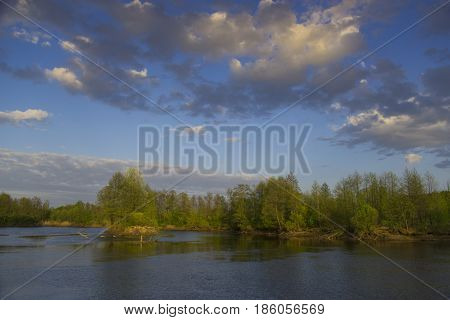 Spring or summer landscape. Water (river) in foreground. Blue sky with clouds in background.