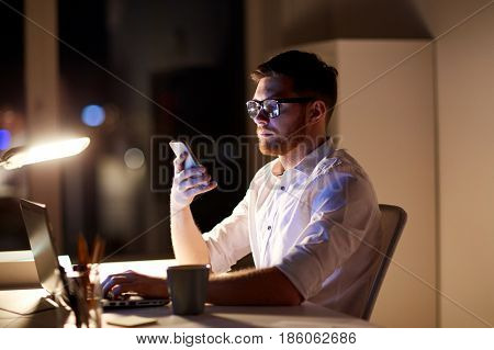 business, overwork, people, deadline and technology concept - businessman in glasses with laptop computer texting on smartphone at night office
