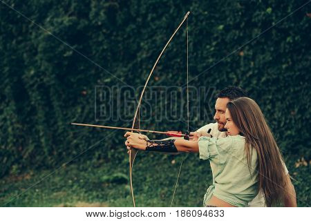 man and pretty woman or cute girl with long hair archers or hunters shooting with bow and arrow on summer day on green natural background. Couple in love. Concentrate and aiming