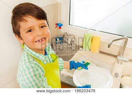 Close-up portrait of smiling kid boy washing dishes with sponge standing next to the sink