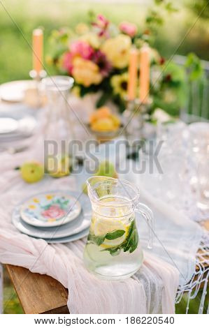 picnic, food, summer, holiday concept - full glass jug of fresh lemonade on festive table, slices of lemon, lime, mint leaves in flagon, bouquet, candles, glasses and plates, fruits, selective focus
