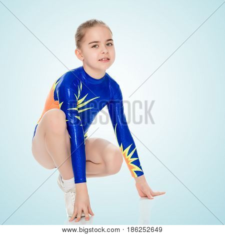 Beautiful Russian girl gymnast younger school age, in blue in a sports swimsuit.The girl squatted down and put her hands on the floor.On the pale blue background.