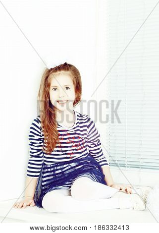 Little girl in blue striped dress and a white bow on her head.She sits at the window and looking straight to the camera.Creative toning of a photograph.