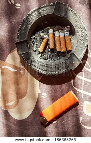 Various cigarette butts in full ashtray and orange plastic lighter on table