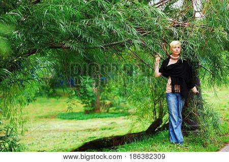 Young pretty blonde girl in blue jeans and a black tippet stands near the willow in the summer.