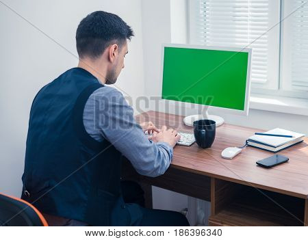 Young man office worker sitting at computer with Chromakey, looking on monitor and typing. Office concept with working environment. Back view. Copyspace on screen